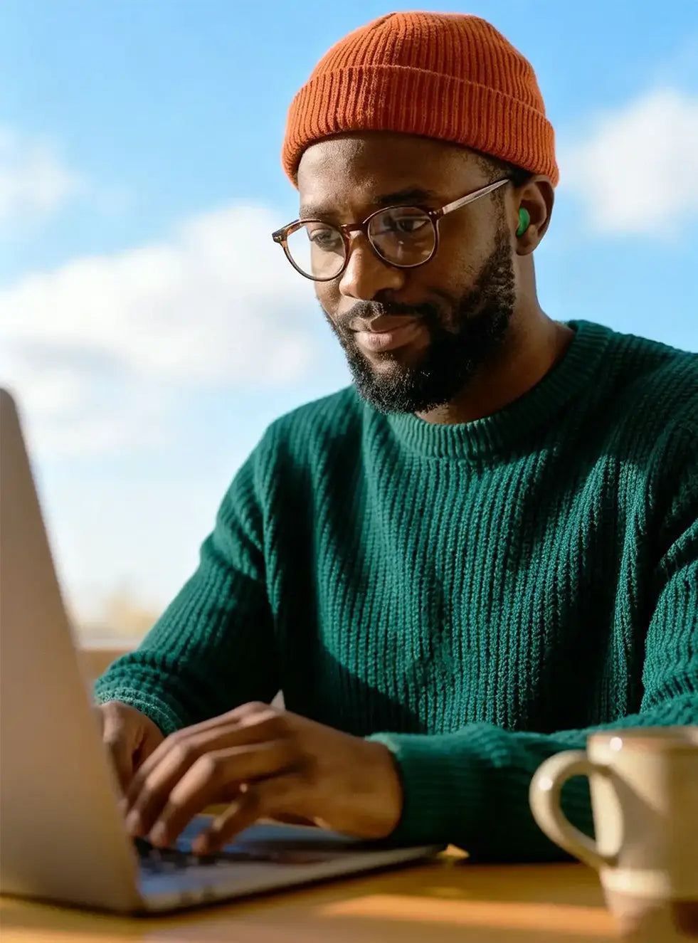 A man wearing an orange beanie and green sweater working on a laptop while wearing Green Soundbuds earplugs for focus.