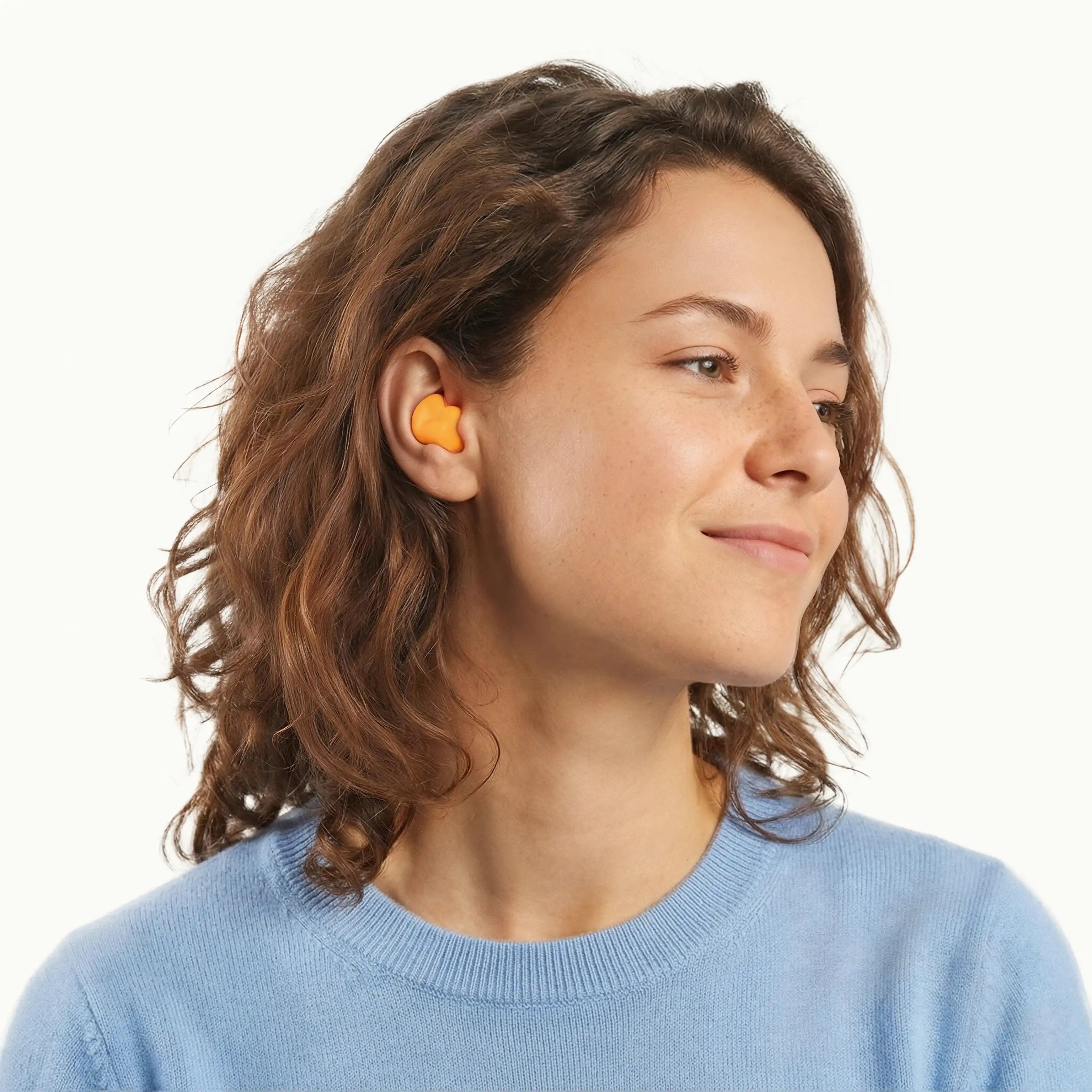 A side profile view of a woman with wavy brown hair wearing a bright orange mouldable silicone Soundbud earplug, shown against a solid white background