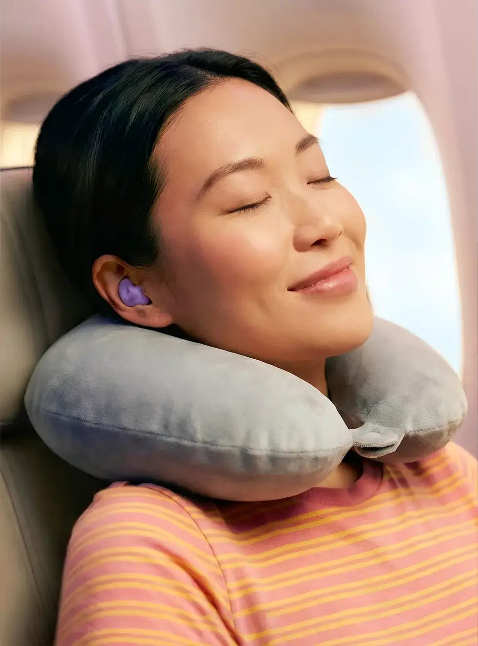A woman sleeps comfortably in an airplane seat using a grey travel neck pillow and purple Happy Sleep earplugs for noise reduction.